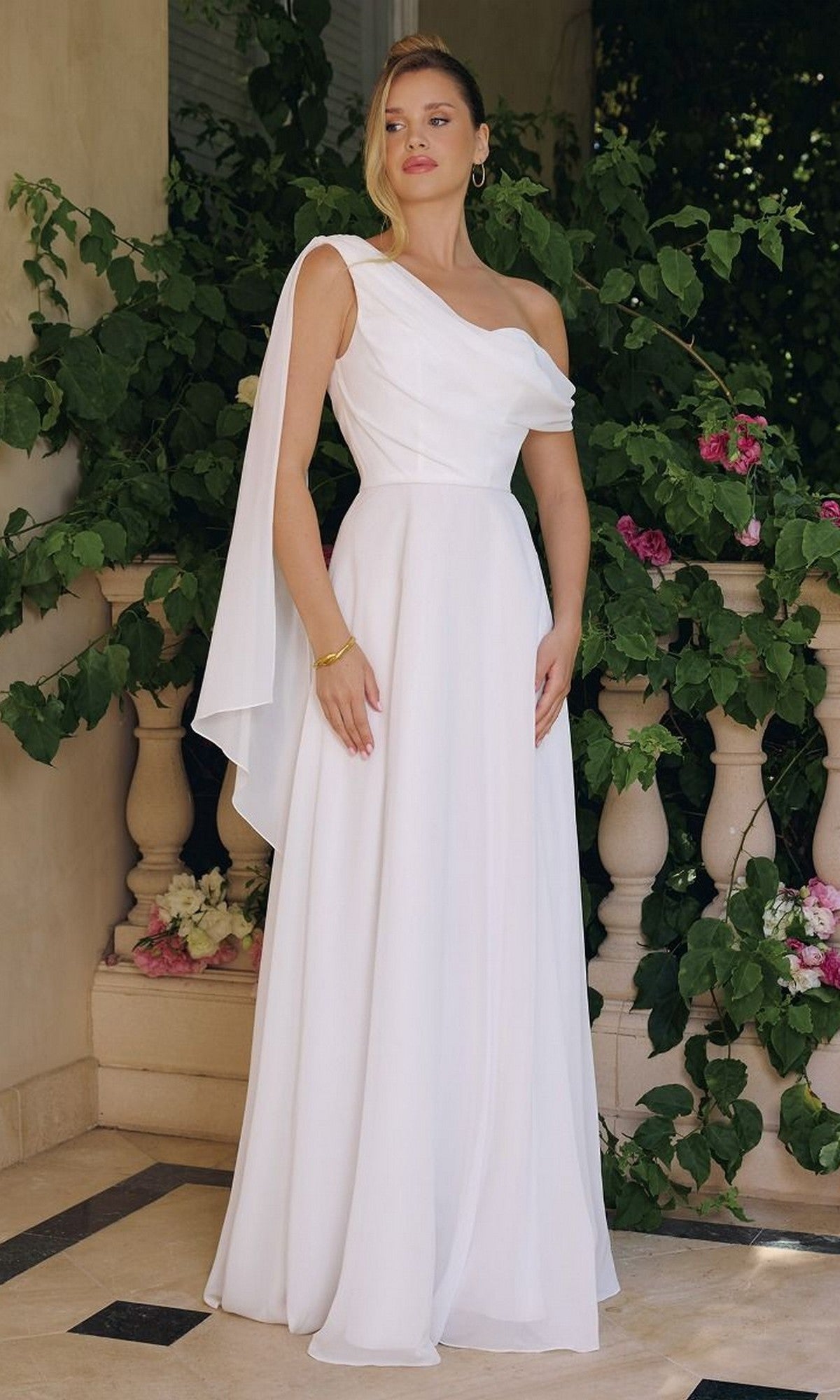 A woman in the Asymmetrical Off-White Bridal Gown: Amoris T716W, featuring a long chiffon skirt, stands on a tiled patio before greenery and a stone railing.
